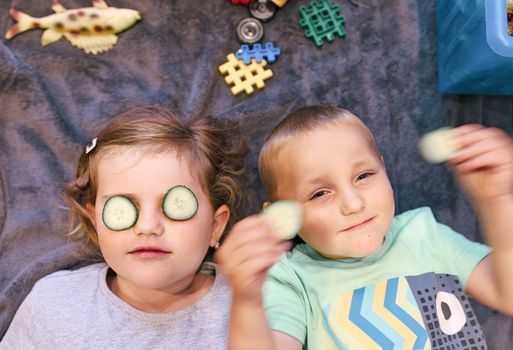 Funny small girl with piece of cucumber on their eyes like a mask with his brother, beauty and health concept, indoor closeup portrait