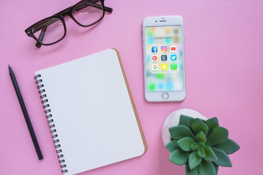 BANGKOK, THAILAND - NOVEMBER 28, 2016:  Group of Popular Social networks icons showing on Apple iPhone 6s screen with notebook, eyeglasses and green plant on pink background on pink background, Social media are most popular tool for communication.