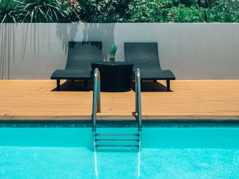 Outdoor swimming pool background minimal style. Grab bars ladder in the blue swimming pool with two black sunbed and green shrubs on grey wall.