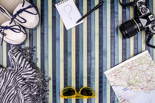 Background composition from above for summer holidays with vintage camera, sunglasses, white shoes, striped foulard, map and notepad with handwritten question mark. Striped wooden background on the light blue.