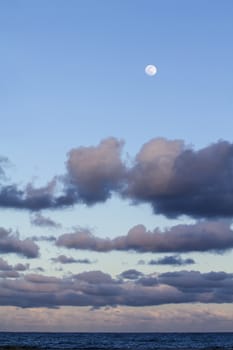 Impressive sky at sunset with a full moon above and layers of clouds tinged with pink that slope down to the horizon over the dark blue sea