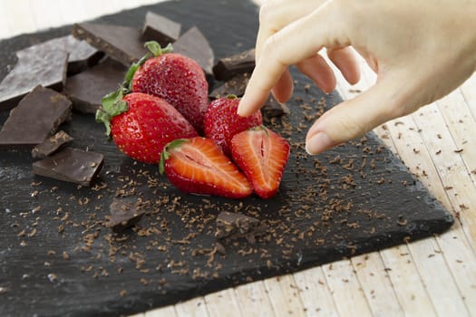 Woman's hand about to take one of the neatly placed strawberries on a slate plate with chopped chocolate and grated around on a light wooden background and selective focus