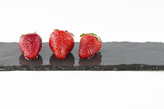 Close-up of three isolated strawberries on a rectangular strip of wet slate on a white background shot in high angle view with selective focus