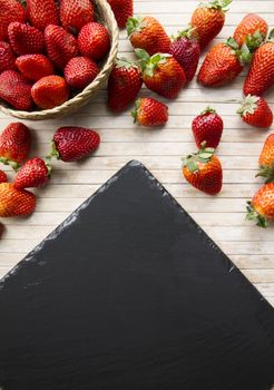 Strawberries copy space with a small basket of strawberries surrounded by strawberries scattered on a wooden bottom and a plate of slate taken from above