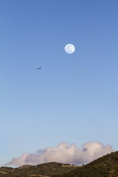 Full moon in the twilight time in the clear blue sky with a cloud resting on the hill below and a peregrine falcon flying