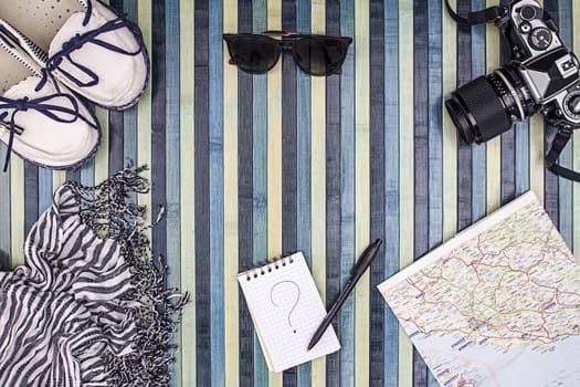 Background composition from above for summer holidays with vintage camera, sunglasses, white shoes, striped foulard, map and notepad with handwritten question mark. Striped wooden background on the light blue.