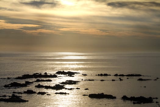 Blue sky at dawn covered by strange cloud formations on a flat sea with black rocks that stand out in the reflection of the sun on the water