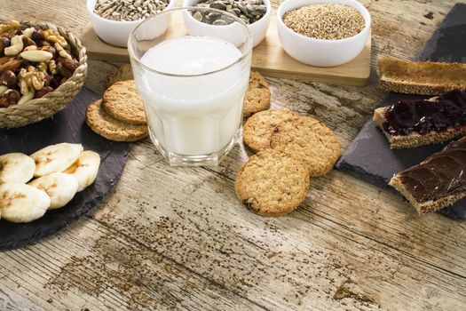 Wooden table set for a sweet vegan breakfast with rice drink, sliced banana, mixed dried fruit, vegan biscuits, slices of bread with jam and cream of veg hazelnuts, pumpkin seeds, sesame and sunflower seeds