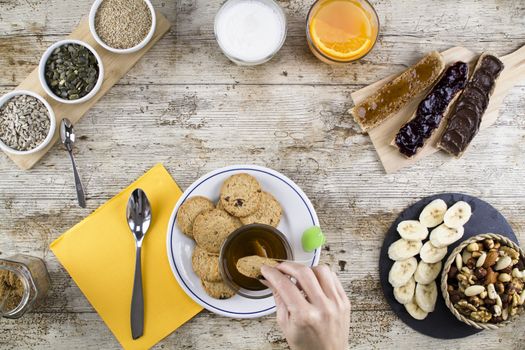 A woman's hand soaking the biscuit in tea at a wooden table set for a sweet vegan breakfast shot from above with sliced banana, mixed dried fruit, vegan biscuits, slices of homemade bread, mixed seeds