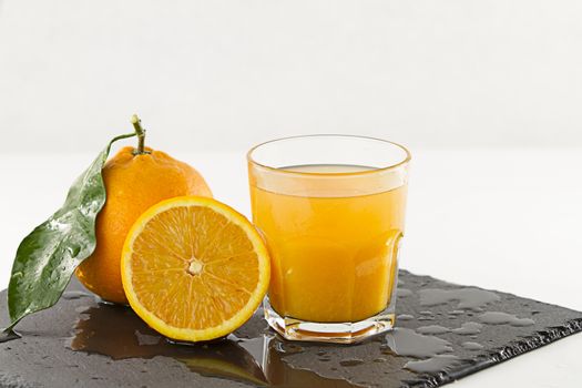 An inviting glass full of orange juice, an half orange and a whole one with leaf on a wet square slate plate onwhite background