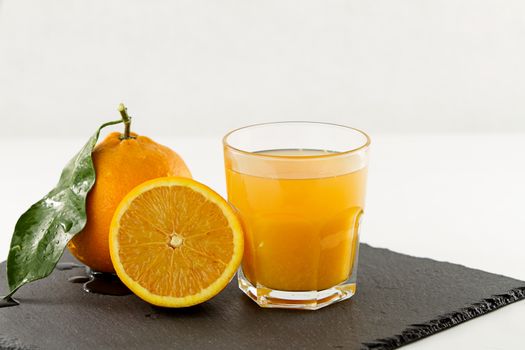 An inviting glass full of orange juice, a half orange and a whole one with leaf on a square slate plate onwhite background