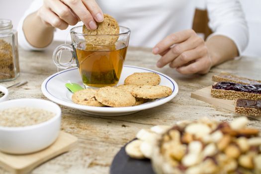 A woman's hand on the table in a wooden table set for a sweet vegan breakfast with a slice of banana, vegan biscuits, mixed seeds, homemade bread