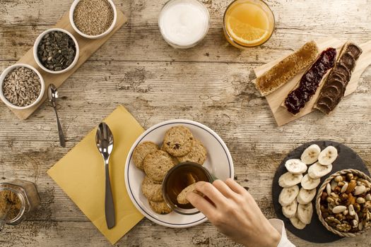 A woman's hand soaking the biscuit in tea at a wooden table set for a sweet vegan breakfast shot from above with sliced banana, mixed dried fruit, vegan biscuits, slices of homemade bread, mixed seeds