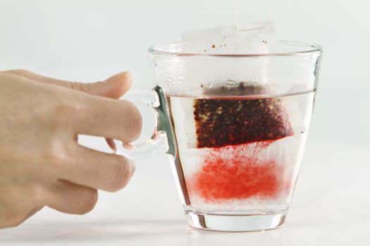 Close-up of a woman hand dipping a sachet of Hibiscus Tea in a glass cup full of water with beautiful red-colored effects in the transparency of water