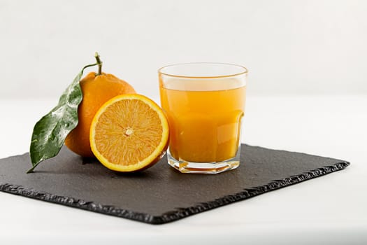 An inviting glass full of orange juice, a half orange and a whole one with leaf on a square slate plate onwhite background
