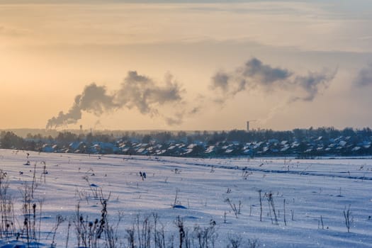 Snowy field on a frosty winter day and a city with smoke from pipes on the horizon