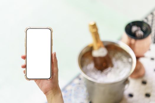 Woman hand holding blank white screen mobile phone with champagne bottle in ice bucket and two glasses near jacuzzi pool.