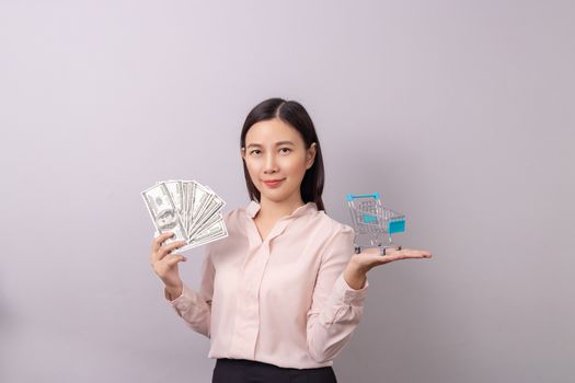 retail commercial business concept, Asian beautiful woman holding banknote money in hand and shopping cart in another hand isolated on grey background