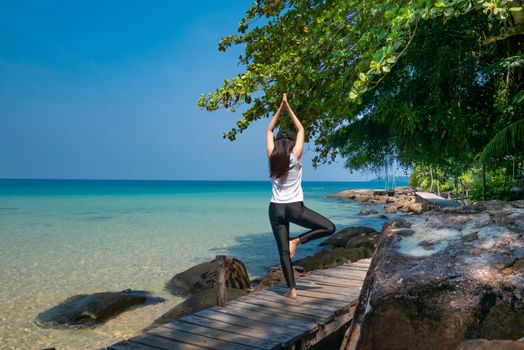 a woman standing on one leg while practicing yoga on wooden bridge over the sea during summer vacation. attractive young Asian woman standing in yoga pose