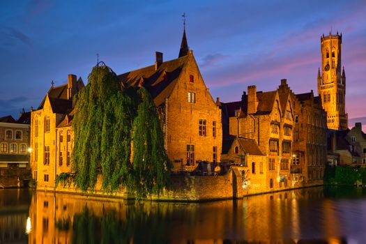 Famous view of Bruges tourist landmark attraction - Rozenhoedkaai canal with Belfry and old houses along canal with tree in the night. Brugge, Belgium