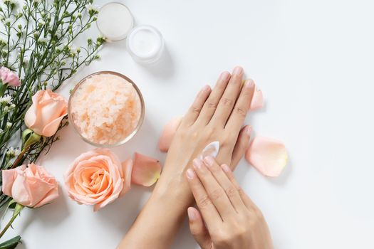 natural skincare concept. woman apply white cream on her hands on white background with jar of cosmetic cream, salt spa scrub ,rose and  white flowers with copy space