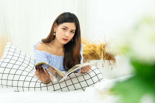 beautiful Asian woman lying on white cozy bed reading a book enjoys of rest at comfort home