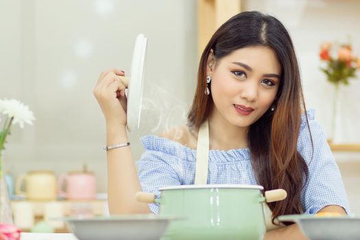 beautiful Asian woman sit at the table in the kitchen, open the lid of the soup pot with the heat vapor floating up.
