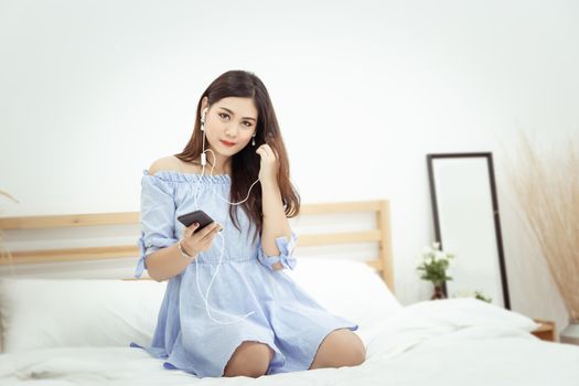 A beautiful Asian woman with headphone relaxing at home , sitting on the bed listening to music from application on mobile phone