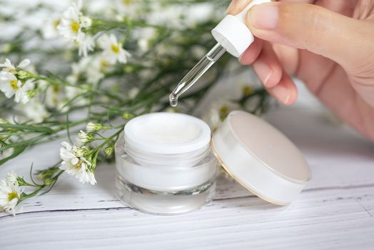 organic natural skincare concept. open blank cosmetic cream jar with white cream texture inside and women hand holding glass dropper for dermatologist testing decorate with white flower at background