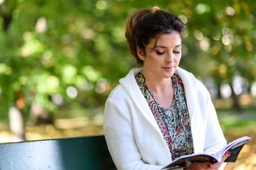 Beautiful young woman sitting on a bench in the park and reading a book on a sunny autumn day.