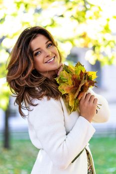 Portrait of a beautiful and lovely young woman smiling and holding fallen leaves on a blurred background of the autumn park on a sunny day.