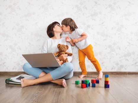 Mom and son at home. Mother works remotely with laptop and kisses her kid. Freelance work in same time with raising children.