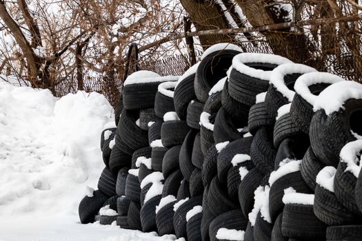 Auto tires are stacked along a chain link fence in the winter, with snow covering parts of them.