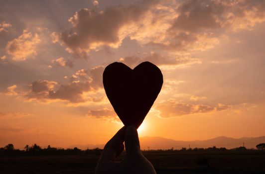 Silhouette of the heart shaped cardboard in hand and the sun shining through the heart on sunset background. Concepts of love and valentines day.
