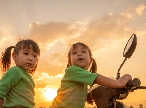 Cute little child girl sitting on motorcycle with her sister over sunset sky background.