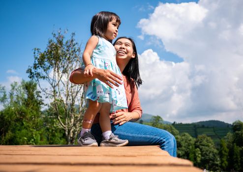 Happy mother sitting with her daughter on wooden ground over trees  with mountains background under blue sky in summer day.