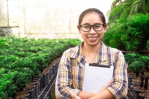 Asian gardener woman standing with tablet in seedling nursery and smile to camera. Technological and agricultural concepts.