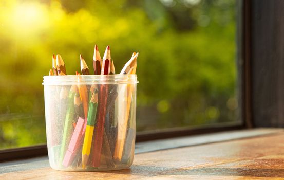 The colored pencils are in a plastic round box placed on a wooden table by the window with the sun shining.