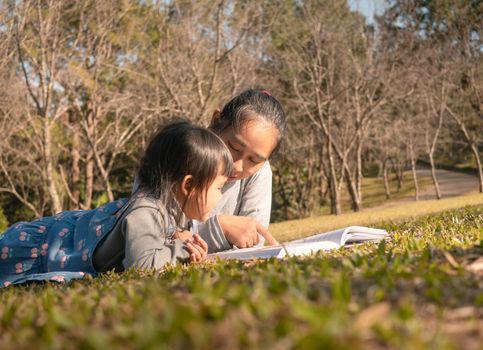 Asian mother reading a book to her daughter in spring garden. Family spending time together on picnic on summer vacation.