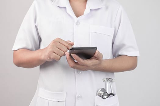 Confident young woman doctor using smartphone with stethoscope in her pocket and stand over grey background.
