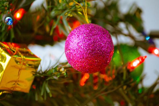 Close up of pink bauble hanging from a decorated Christmas tree with lights presents for new year isolated on white background.