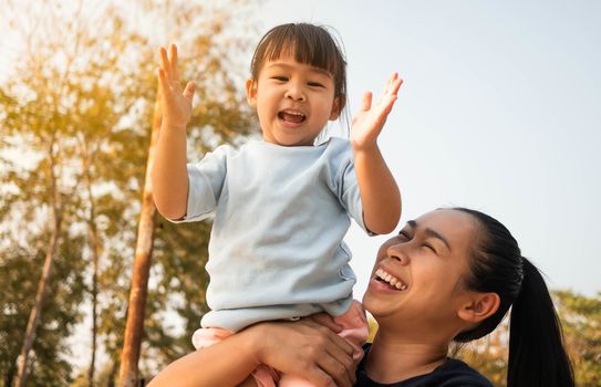 Attractive Asian mother carrying her daughter on shoulder outdoors in the park. Happy family spending free time together.