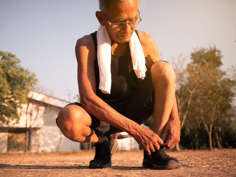 Asian senior man tying his laces of running shoes to prepare jogging in the park for good health. Healthcare concept.
