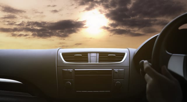 Driver's hand on the black steering wheel with dashboard inside of a car and see the view of the sunset sky through the windshield. Vintage tone.