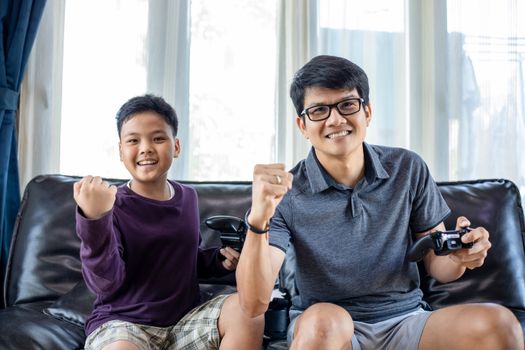 Asian father and son enjoy playing video games together with video joystick with excite and very fun in the living room at home