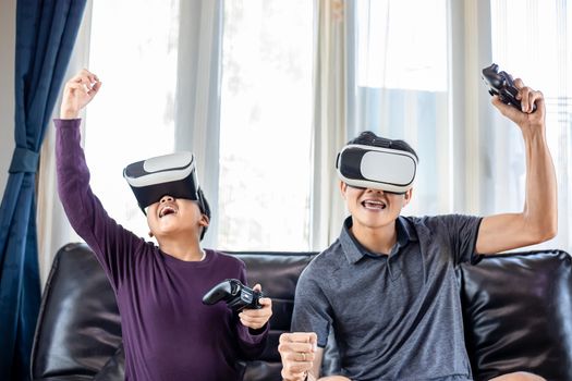 Asian father and son enjoy playing video games together with video joystick and Virtual Reality Glasses with exciting and very fun in the living room at home