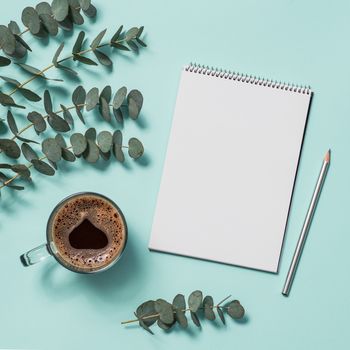 Blank paper notebook with pencil and coffee cup. Empty paper sketchbook with pincil and eucalyptus branches on blue background. Top view or flat lay. Copy space for text or design.