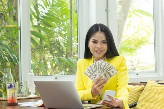 portrait of cheerful young Asian woman holding money banknotes and mobile phone with smile face in front of computer notebook at workplace