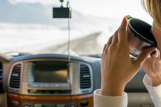 The girl in the passenger seat of the car drinking hot tea from a mug of a thermos, looking on the mountain winter road outside the car window