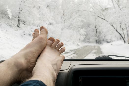 bare human feet on the windshield panel of the car on the background of snowy winter forest landscape and road